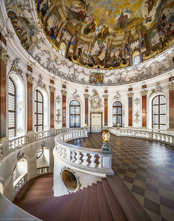 Interior of a beautiful Baroque building with a grand staircase and elaborate painted dome, showcasing beautiful architecture.