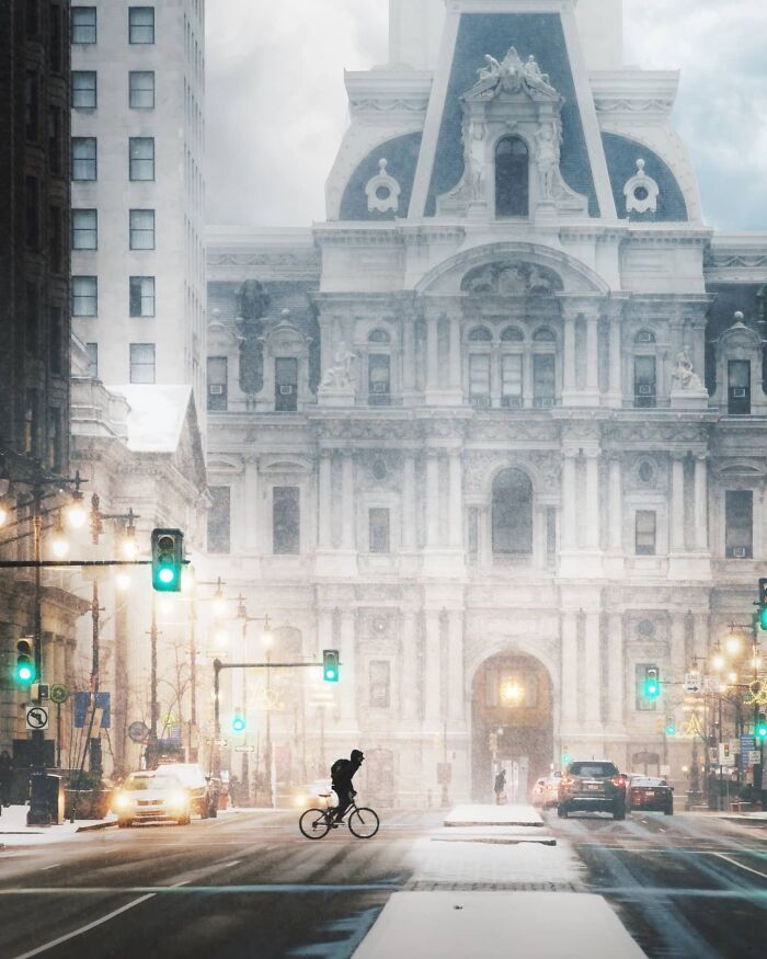 Snow falling on beautiful architecture in a city, with a person riding a bicycle past the grand building, creating an almost unreal scene.