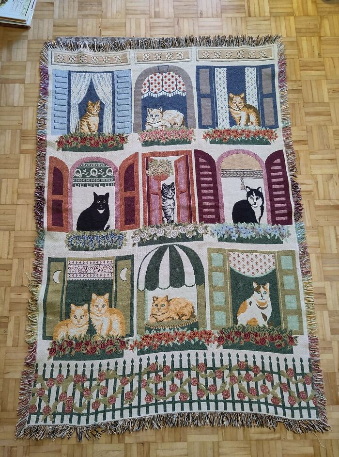 Decorative thrift store tapestry featuring various cats in window scenes with floral accents on a wooden floor.
