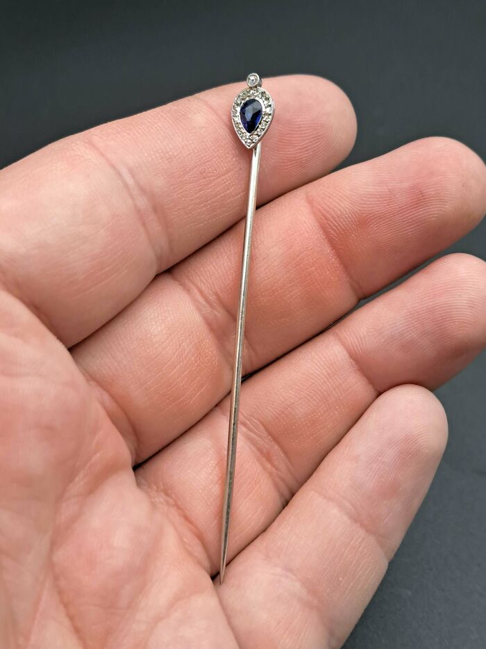 Hand holding a vintage silver stick pin with a teardrop-shaped sapphire surrounded by small diamonds, thrift store find.