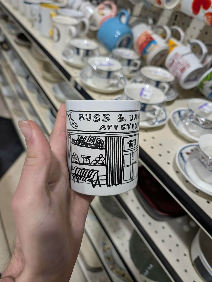 Hand holding a thrift store find mug with a black and white storefront sketch, surrounded by shelves of vintage dishware.