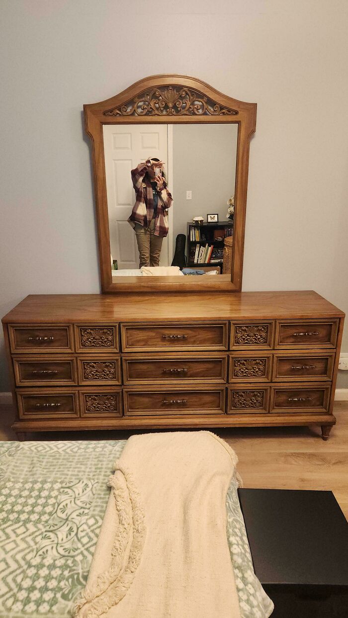 Wooden dresser with ornate mirror, shown as a thrift store find reflecting a person taking a photo in a bedroom.