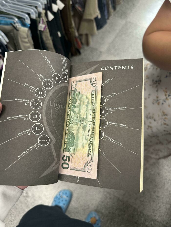Opened book with a $50 bill inside, held in a thrift store environment with clothing racks in the background.