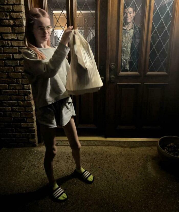 Woman holding a delivery bag outside a door while a delivery driver stands behind a glass panel in a hilarious delivery confirmation pic.