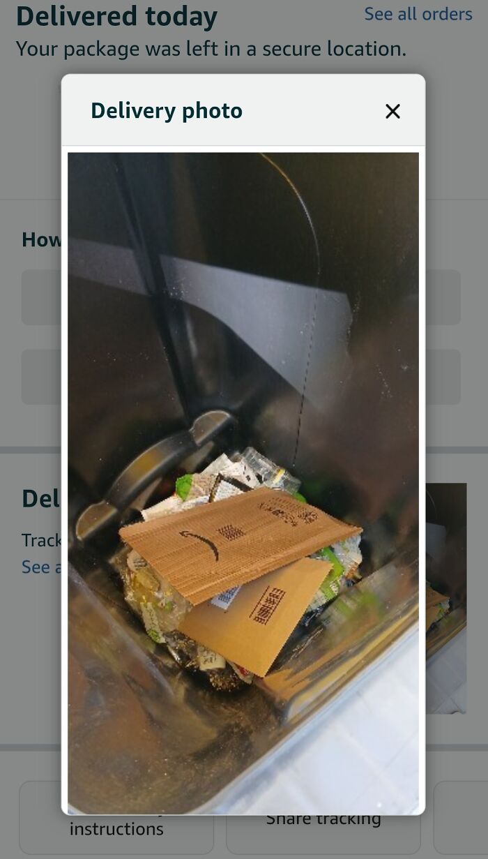 Delivery confirmation pic showing package left in a trash bin with a sad face cardboard, humorous delivery photo.