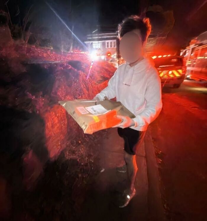 Person holding a package on a dark street with emergency vehicle lights in the background, delivery confirmation pic.