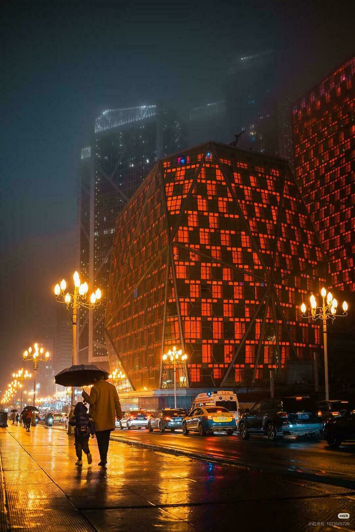 A city street at night with an illuminated, geometrically patterned building that looks like an evil mastermind's lair.