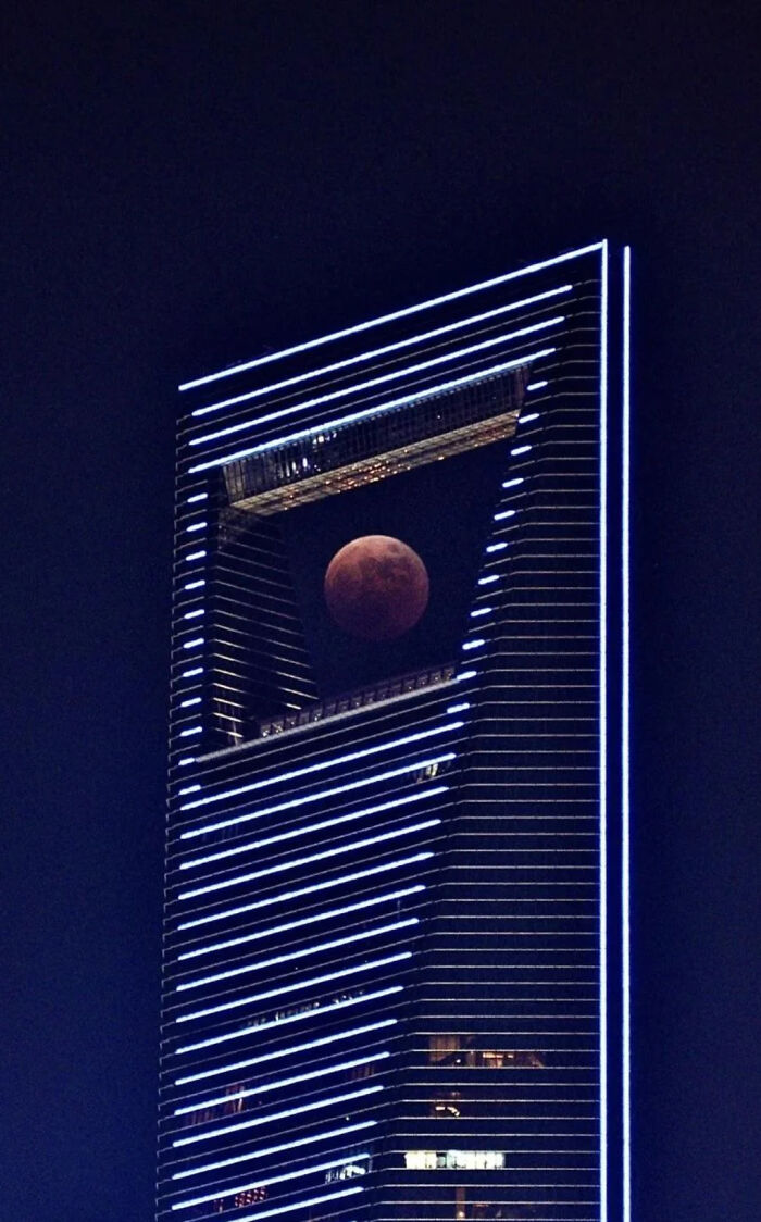 A tall skyscraper with a blood moon perfectly framed in its central opening, evoking an evil mastermind's base.