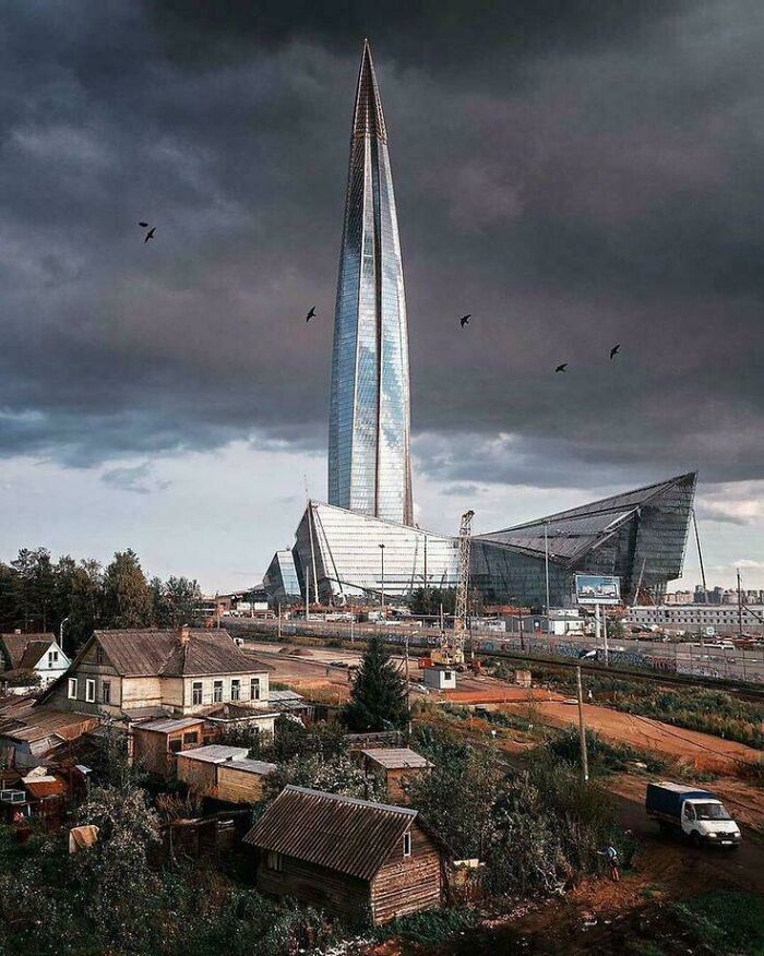 A towering, futuristic skyscraper, a perfect evil mastermind building, under a dramatic cloudy sky, with old houses in the foreground.