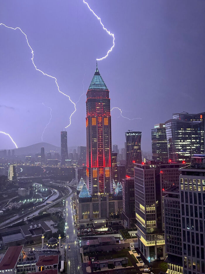 Tall, modern buildings illuminated in red and blue under a stormy sky with dramatic lightning, perfect for an evil mastermind.