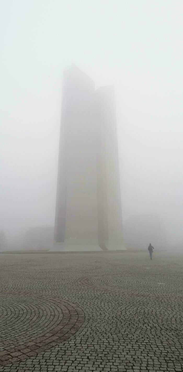 A person walks through thick fog towards tall, ominous buildings, evoking an evil mastermind's plan.