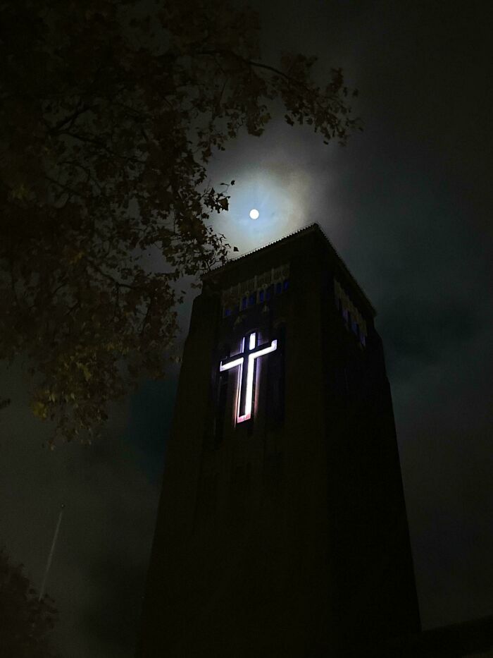 A dark tower with a glowing cross, resembling a building for an evil mastermind, under a full moon.