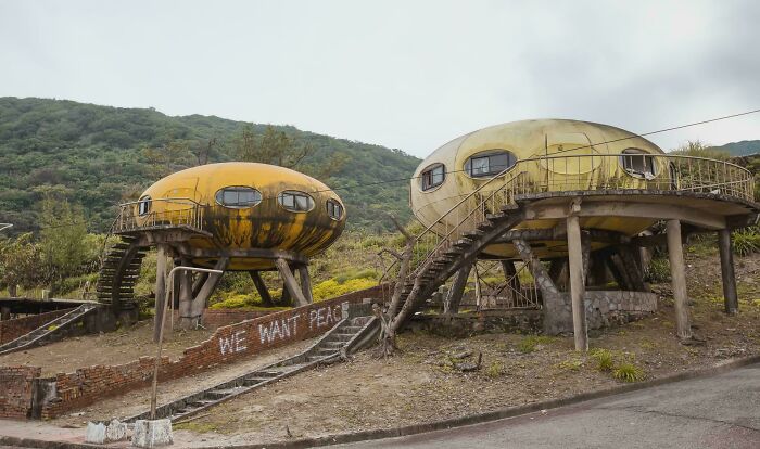 Two abandoned, pod-like evil mastermind buildings, weathered and yellow, set against a green hillside.