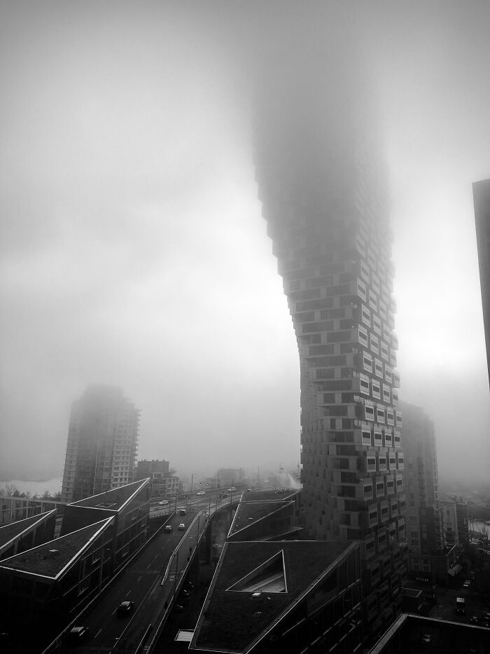 A monochrome view of a curved, modern skyscraper building shrouded in fog, looking like an evil mastermind's headquarters.