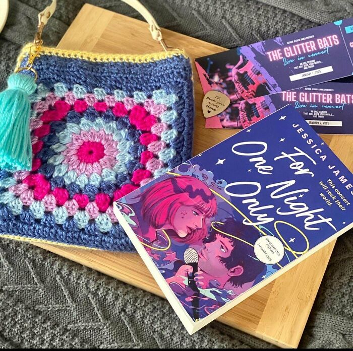A handcrafted crocheted bag in blue and pink granny square pattern, with a book and concert tickets. People crocheted something awesome.