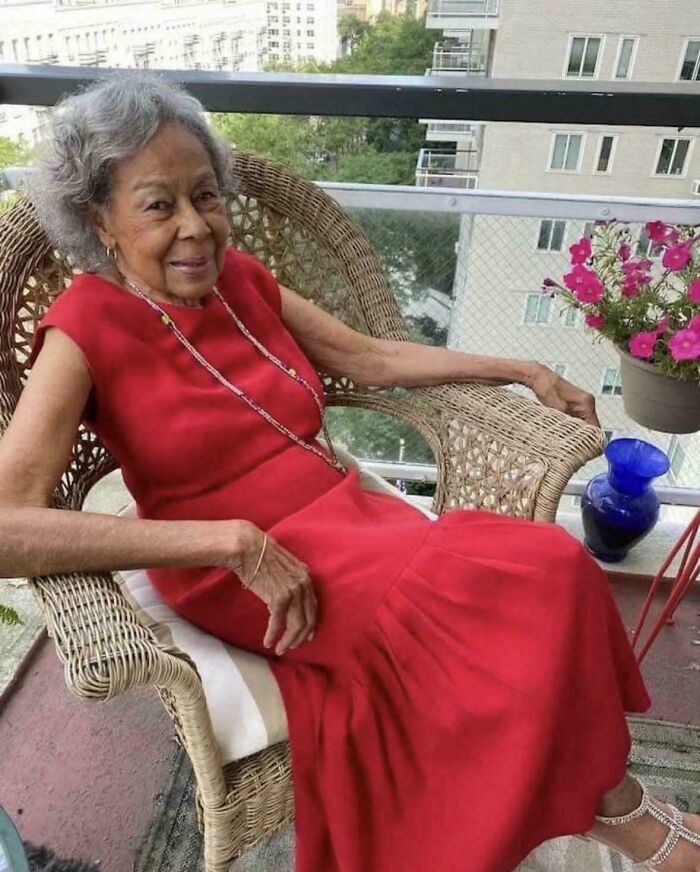 An amazing photo of an elderly woman with grey hair smiling while sitting in a wicker chair on a balcony.