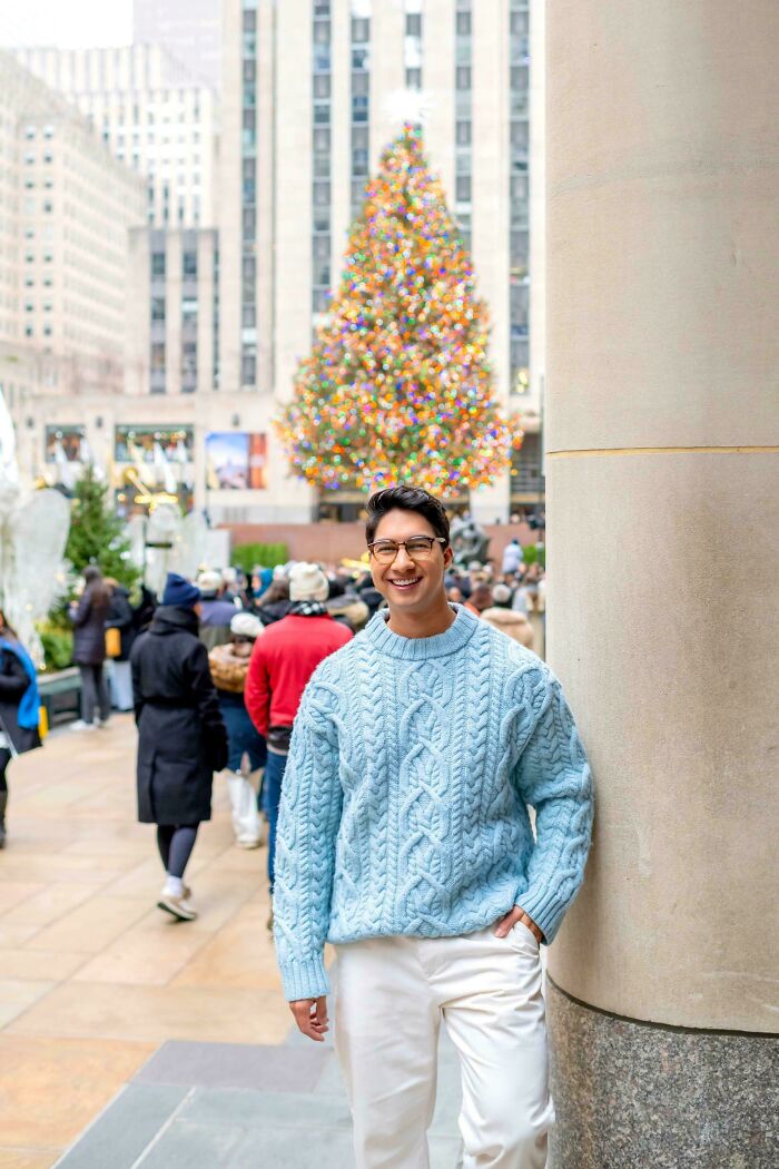 A man in a light blue sweater smiles in front of the Rockefeller Center Christmas tree, showing people not natural on social media.