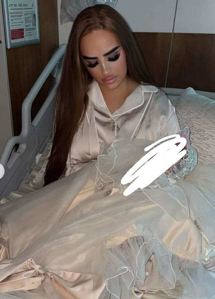 A young woman with long brown hair, heavy makeup, and a satin nightgown sitting in a hospital bed with a blurred object. The image shows people not natural on social media.