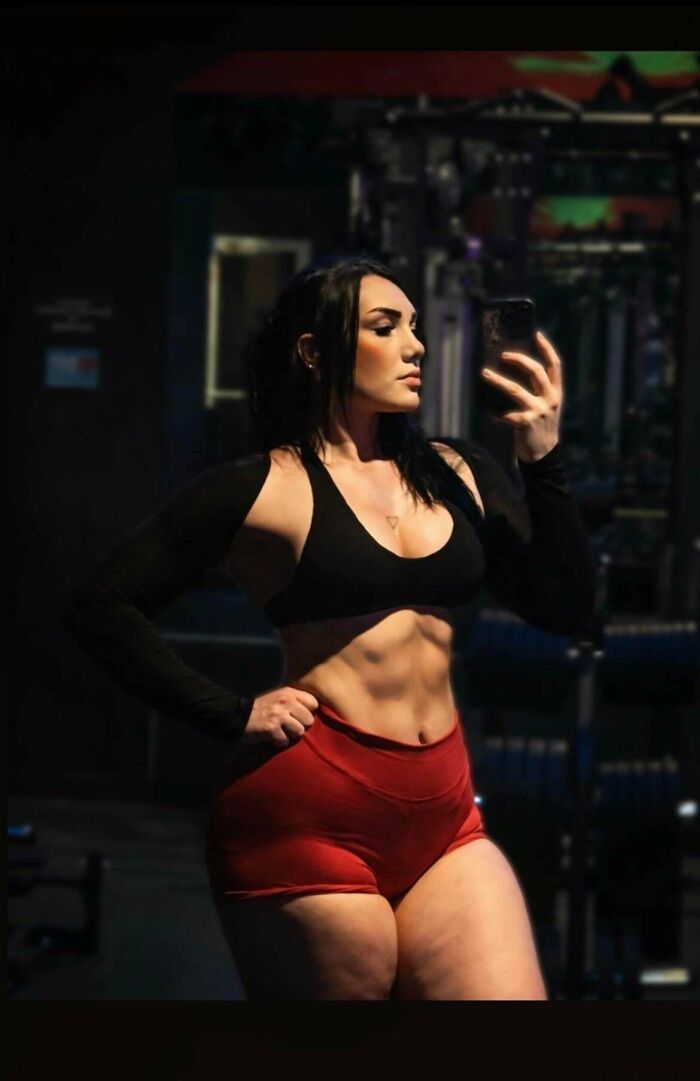 A woman in a sports bra and red shorts poses for a natural social media photo in a gym.