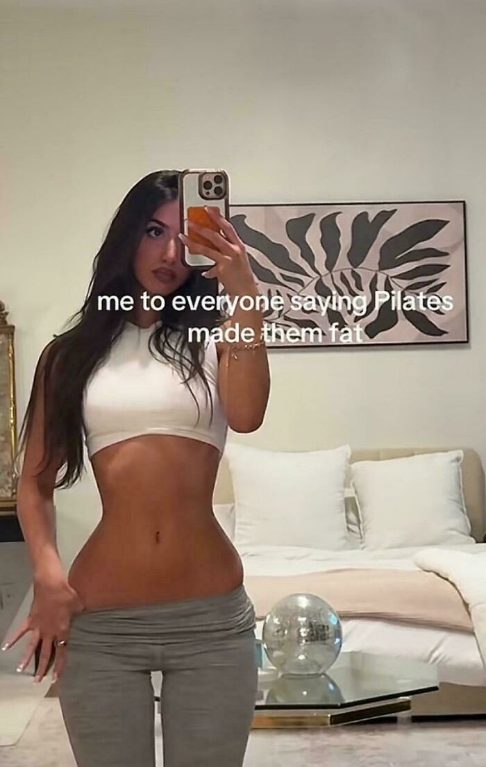 A woman with a cinched waist poses in a mirror, looking natural on social media, wearing a white crop top.