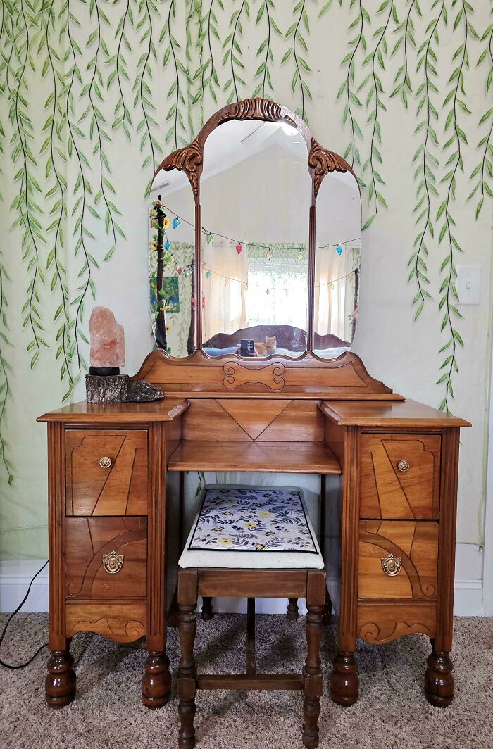 A beautiful antique wooden vanity with a three-panel mirror, a great thrift store jackpot, in a room with leaf-patterned wallpaper.