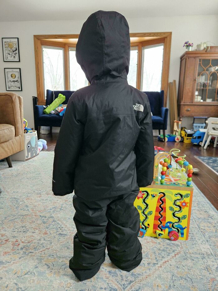 A child in a black snowsuit, a thrift store jackpot find, standing in a living room.