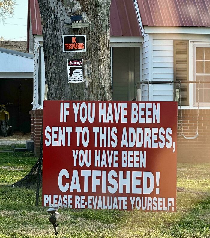 Large red sign in yard warning visitors they have been catfished with other no trespassing signs on a tree behind it.