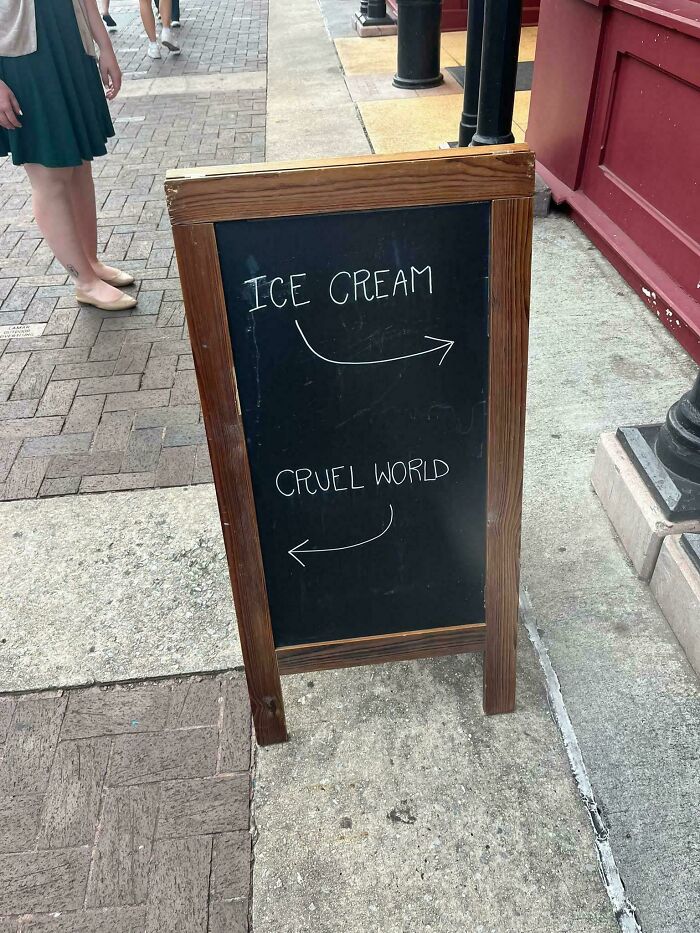 Chalkboard sign outside shop with arrows pointing to ice cream and cruel world, a bizarre and hilarious street sign.