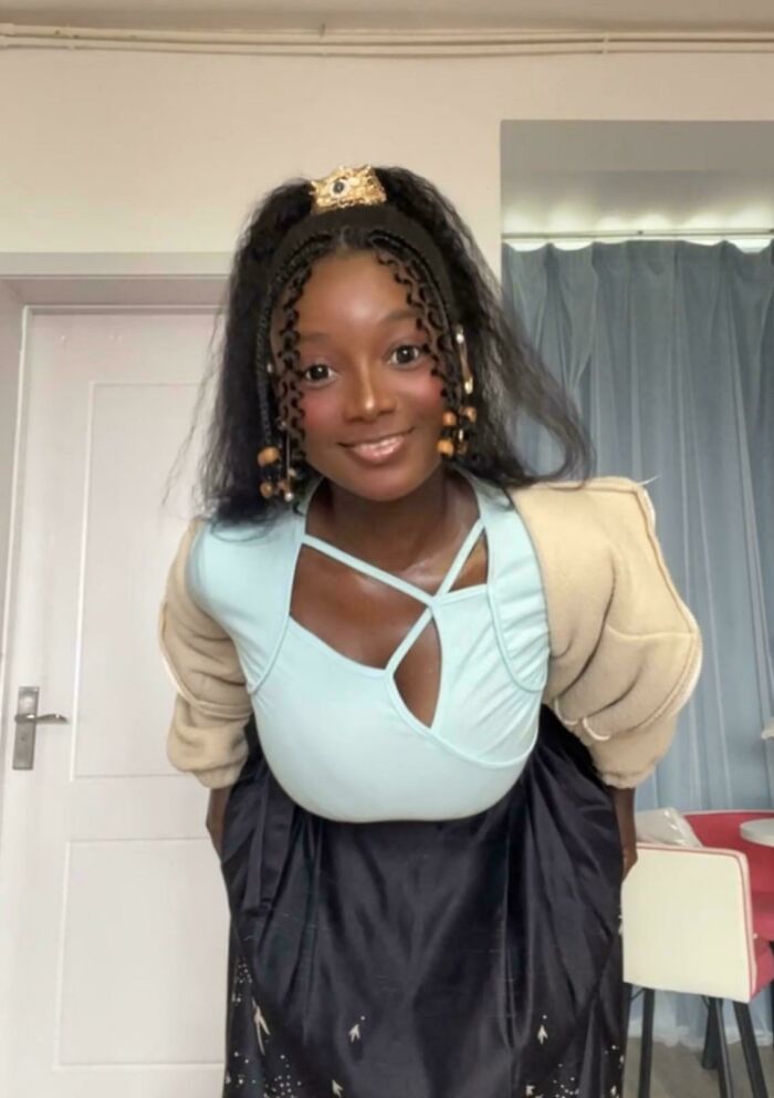 Smiling woman with braided hair and beads, looking natural on social media in a light blue top and black skirt.