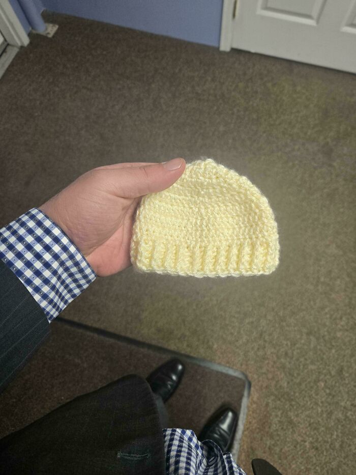 A person in a suit and checkered shirt holding a small, yellow crocheted baby hat. Awesome crocheted item.