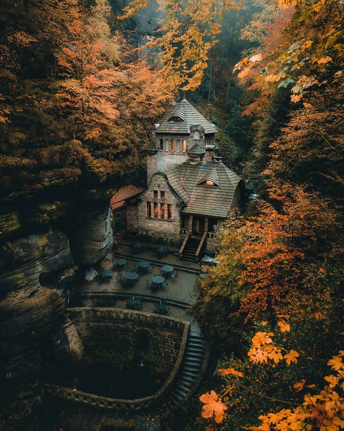 A beautiful example of architecture: a charming stone house nestled in an autumn forest with vibrant orange trees and rocky cliffs.