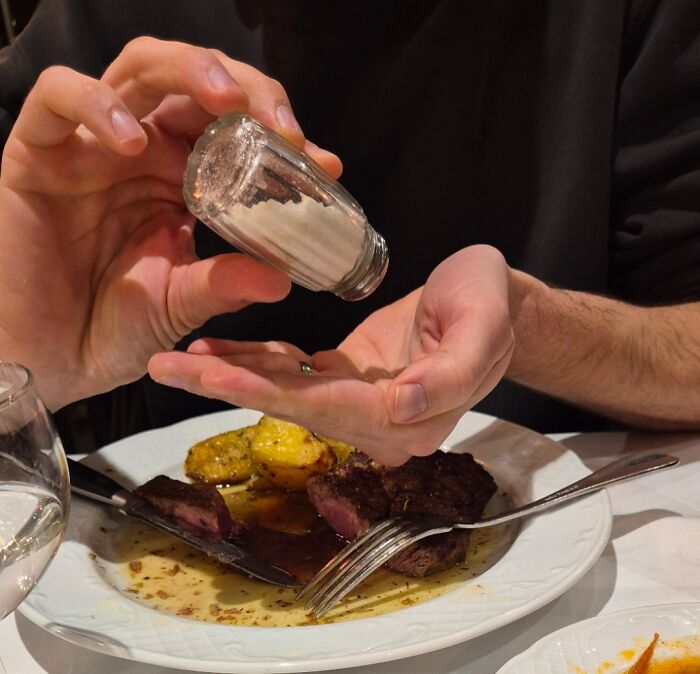 Man seasoning steak with excessive salt directly into his hand, showing typical infuriating examples of men being men.