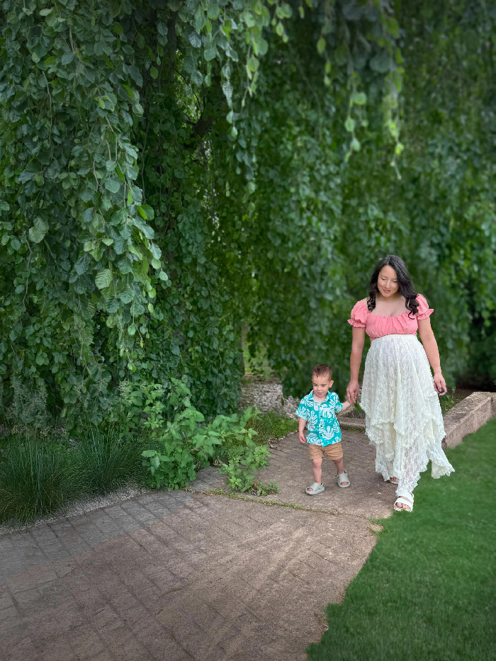 Pregnant woman in lace dress holding toddler's hand on garden path under hanging foliage, maternity photoshoot