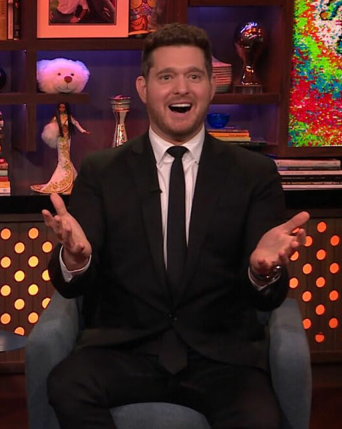 Man in black suit and tie gesturing with hands while seated, in a colorful studio with decorative background elements.