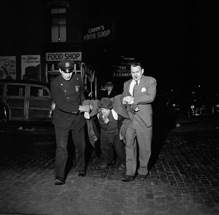Police officer and man assist an intoxicated man on a cobblestone street in a vintage black and white street photo.