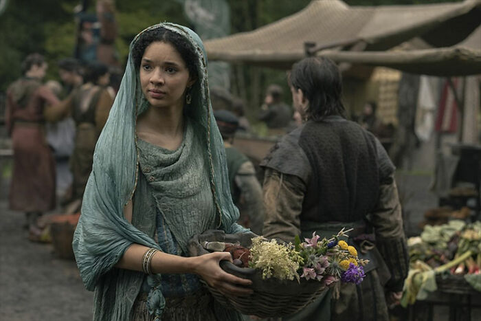 Young woman in medieval attire holding a basket of flowers in a market scene from A Knight Of The Seven Kingdoms cast members. Young woman in medieval attire holding a basket of flowers in a market scene from A Knight Of The Seven Kingdoms cast members.