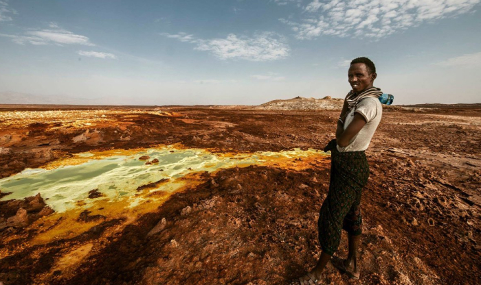 Man standing near a colorful acidic pool in one of the forbidden places on Earth with rugged, barren terrain under a cloudy sky.