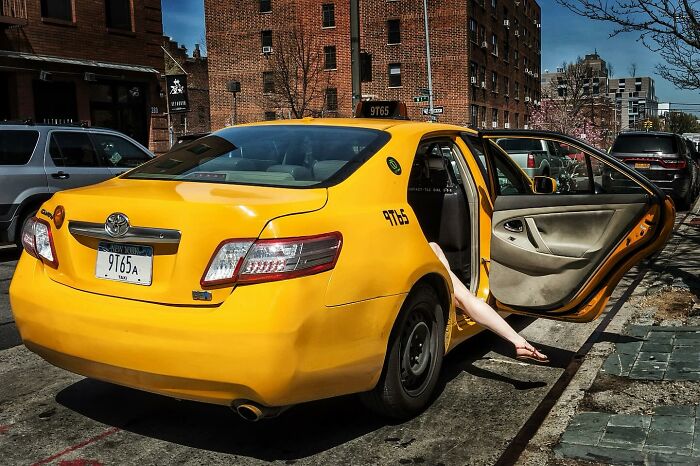 Yellow taxi parked on city street with open door and person’s leg extended, candid street photo capturing human moments.
