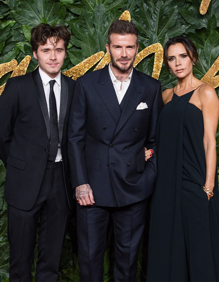 Brooklyn Beckham with Victoria Beckham and David Beckham at an event posing against a leafy, decorative background.