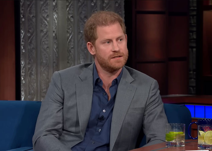 Prince Harry in a navy shirt and gray blazer, seated on a talk show set with drinks on the table nearby.