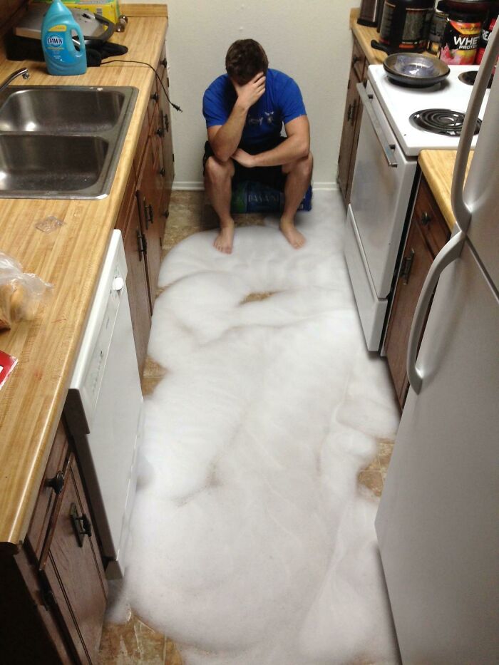Man sitting in kitchen overwhelmed by large foam spill covering the floor, showcasing jerks who frustrate their roommates.