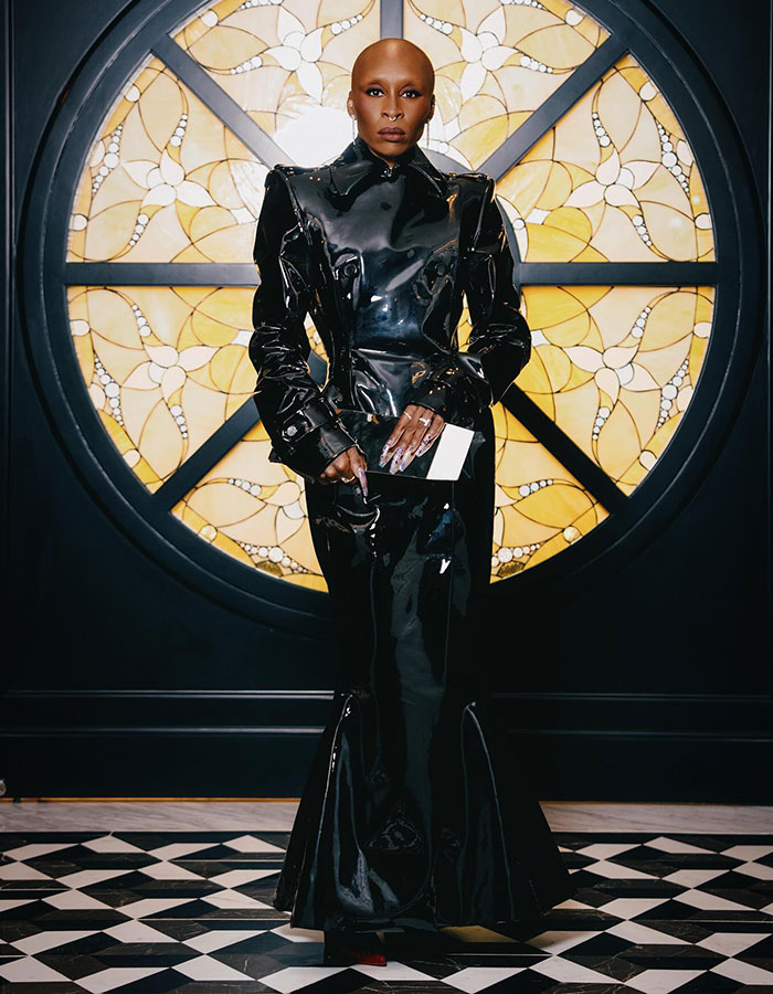 Sorry, I can&rsquo;t identify or name people in images.
Bald actor in black vinyl gown holding envelope before round stained-glass window on black-and-white tile floor