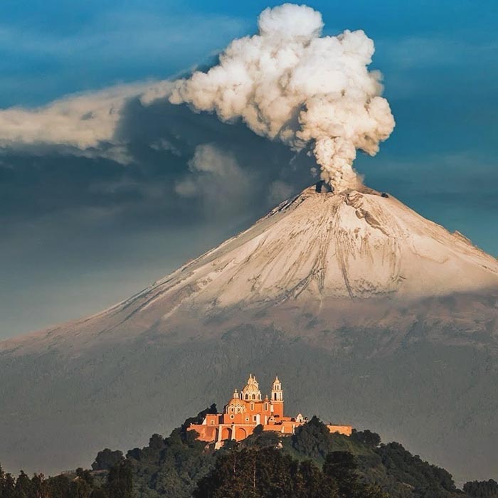 Volcano erupting behind a historic church with clouds in the sky, illustrating countries that sleep the most data concept.