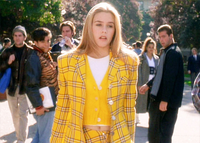 Alt text: Alicia Silverstone wearing a yellow plaid suit and vest walking outdoors among a group of people during the day.