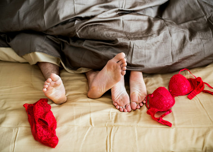 Feet of a couple under bed covers with red lingerie nearby, illustrating challenges faced by an intimacy surrogate on the job.