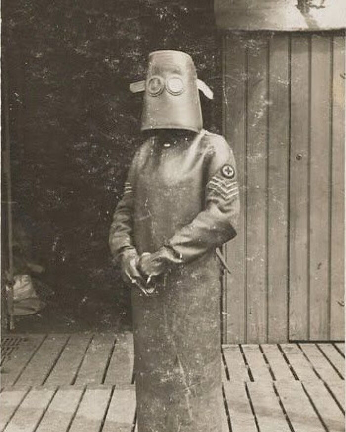 Vintage black and white photo of a person in a full odd and creepy protective suit with a bucket-like helmet.