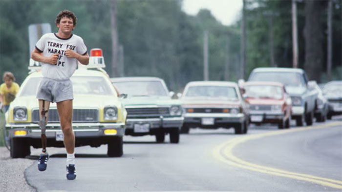 Runner with prosthetic leg wearing Terry Fox Marathon of Hope shirt running on road with cars following, representing impactful country stories.