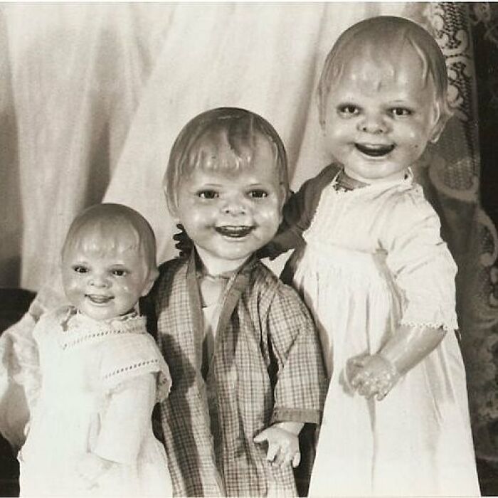 Three old black and white creepy dolls with unsettling smiles and worn clothing in a vintage eerie setting.