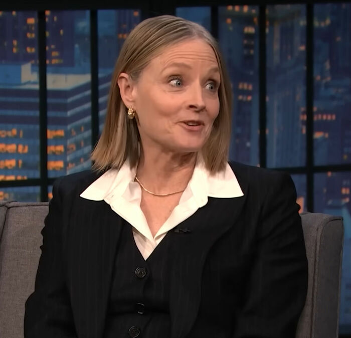 Jodie Foster speaking in a TV interview, wearing a black suit and white shirt, discussing Hollywood predators and her childhood.