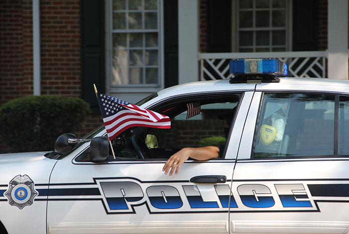 Police car parked with an American flag on the window and a person&rsquo;s arm resting outside the door.